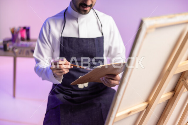 استخدام ريشة الرسم والتلوين، ممارسة الهوايات المفضلة، الجلوس أمام اللوحة، بورتريه مقرب لرسام عربي خليجي سعودي يرتدي الثوب التقليدي ومريلة الرسم، أنشطة ترفيهية فنية ممتعة، خلفية ملونة