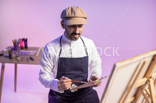 ممارسة الهوايات المفضلة، الجلوس أمام اللوحة، بورتريه مقرب لرسام عربي خليجي سعودي يرتدي الثوب التقليدي ومريلة الرسم، استخدام ريشة الرسم والتلوين، أنشطة ترفيهية فنية ممتعة، خلفية ملونة