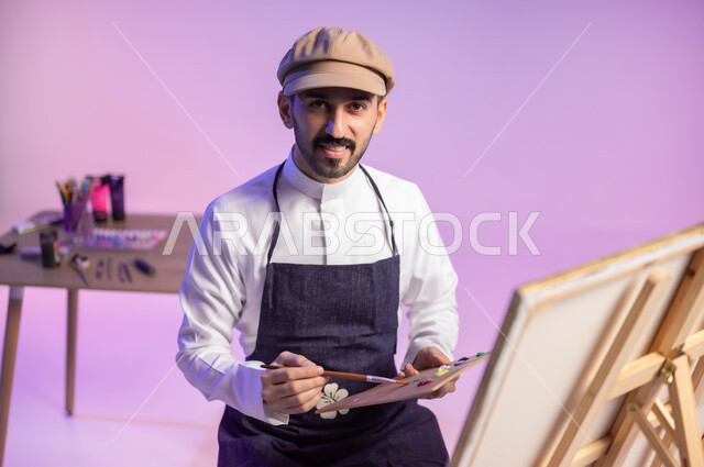 Looking at the camera with expressions of happiness and accomplishment, sitting in front of the painting, a close-up portrait of a Saudi Gulf Arab painter wearing a traditional dress and a drawing apron, using a brush to draw and paint, fun artistic recreational activities, practicing favorite hobbies, a colorful background