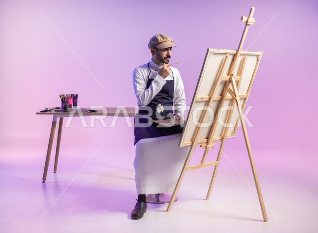 الجلوس أمام اللوحة بتعابير التركيز والتأمل والتفكير، بورتريه لرسام عربي خليجي سعودي يرتدي الثوب التقليدي ومريلة الرسم، استخدام ريشة الرسم والتلوين، أنشطة ترفيهية فنية ممتعة، ممارسة الهوايات المفضلة، الجسم بالطول الكامل، خلفية ملونة