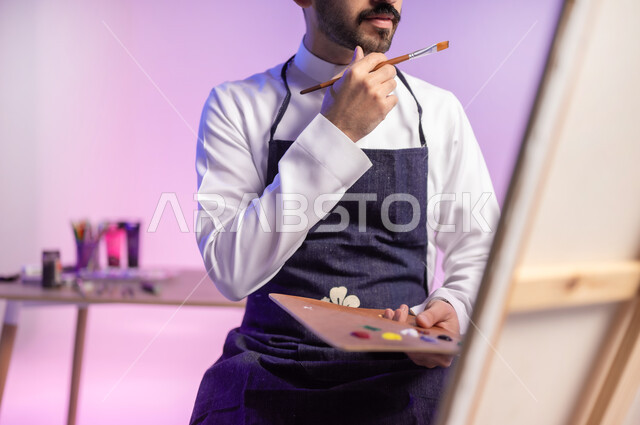 استخدام ريشة الرسم والتلوين، أنشطة ترفيهية فنية ممتعة، الجلوس أمام اللوحة بتعابير التركيز والتأمل والتفكير، بورتريه مقرب لرسام عربي خليجي سعودي يرتدي الثوب التقليدي ومريلة الرسم، ممارسة الهوايات المفضلة، خلفية ملونة