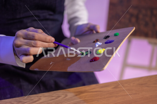 استعمال اللون الأخضر من خلال استخدام ريشة الرسم والتلوين، الجلوس خلف الطاولة وبجانبه لوحة معالم وعلم المملكة، ممارسة هواية الفنون اليدوية، بورتريه مقرب لرسام عربي خليجي سعودي يرتدي الثوب التقليدي ومريلة الرسم، إحياء ذكرى اليوم الوطني، خلفية ملونة