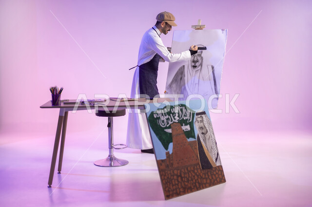 ممارسة هواية الفنون اليدوية، القيام برسم لوحة وطنية، الوقوف أمام لوحة معالم وعلم المملكة، بورتريه لرسام عربي خليجي سعودي يرتدي الثوب التقليدي ومريلة الرسم، استخدام ريشة الرسم والتلوين، إحياء ذكرى اليوم الوطني، الجسم بالطول الكامل، خلفية ملونة