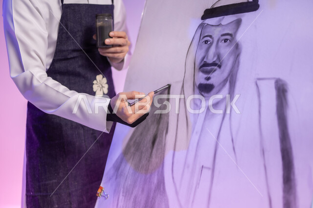 أنشطة ترفيهية فنية ممتعة، الوقوف أمام لوحة معالم وعلم المملكة، بورتريه مقرب لرسام عربي خليجي سعودي يرتدي الثوب التقليدي ومريلة الرسم يقوم برسم لوحة وطنية، استخدام ريشة الرسم والتلوين، إحياء ذكرى اليوم الوطني، ممارسة هواية الفنون اليدوية، خلفية ملونة