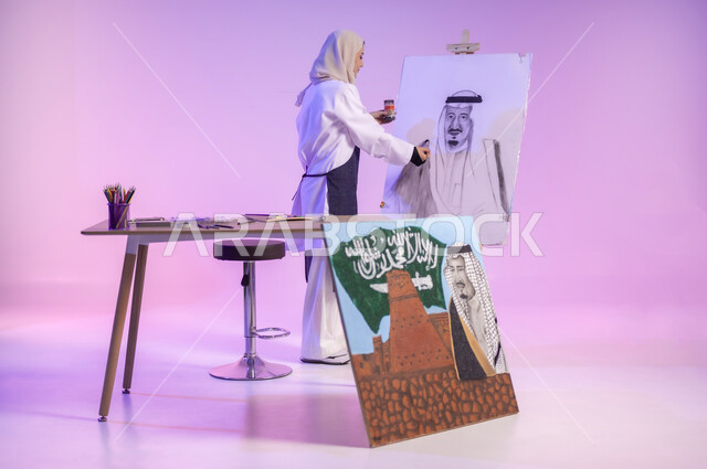 أعياد ومناسبات وطنية، استذكار اليوم الوطني، حب الوطن والإنتماء إليه، استخدام ريشة الرسم و التلوين، الوقوف خلف الطاولة أمام لوحة معالم وعلم المملكة، بورتريه لرسامة عربية خليجية سعودية ترتدي اللباس الكاجوال ومريلة الرسم، الجسم بالطول الكامل، خلفية ملونة