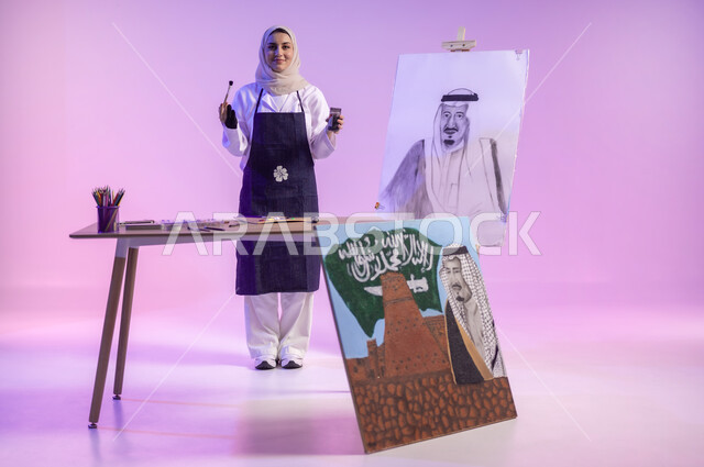 ممارسة مهارات مسلية وممتعة، استخدام ريشة الرسم و التلوين، الوقوف خلف الطاولة أمام لوحة معالم وعلم المملكة، بورتريه لرسامة عربية خليجية سعودية ترتدي اللباس الكاجوال ومريلة الرسم، استذكار اليوم الوطني، حب الوطن والإنتماء إليه، الجسم بالطول الكامل، خلفية ملونة