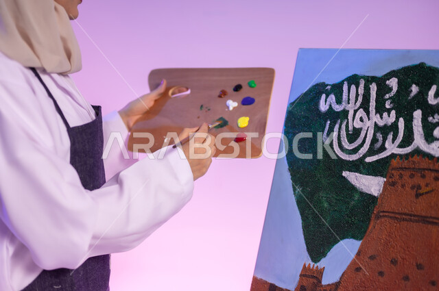 إحياء ذكرى اليوم الوطني السعودي، الأعياد والمناسبات الوطنية، ممارسة مهنة الرسم، الوقوف أمام لوحة معالم وعلم المملكة، بورتريه مقرب لرسامة عربية خليجية سعودية ترتدي اللباس الكاجوال ومريلة الرسم، استخدام أدوات ومعدات التلوين، خلفية ملونة