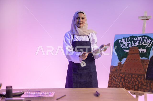 النظر للكاميرا بإيماءات السعادة، هواية الفنون اليدوية والرسم، الوقوف خلف الطاولة وبجانبها لوحة معالم وعلم المملكة، بورتريه مقرب لرسامة عربية خليجية سعودية ترتدي اللباس الكاجوال ومريلة الرسم، استخدام أدوات ومعدات التلوين، استذكار اليوم الوطني السعودي، خلفية ملونة