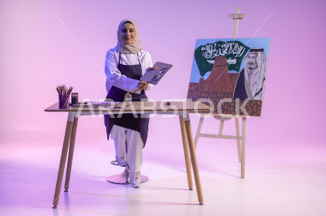أنشطة ترفيهية فنية، استخدام أدوات ومعدات التلوين، الجلوس خلف الطاولة وبجانبها لوحة معالم وعلم المملكة، بورتريه لرسامة عربية خليجية سعودية ترتدي اللباس الكاجوال ومريلة الرسم، استذكار اليوم الوطني، النظر للكاميرا بإيماءات السعادة، الجسم بالطول الكامل، خلفية ملونة