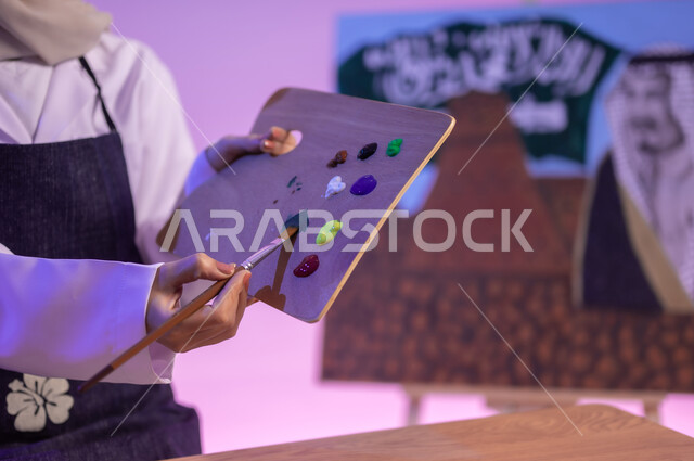 استخدام أدوات ومعدات التلوين، دمج الألوان مع بعضها وتركيب لون جديد، الجلوس خلف الطاولة وبجانبها لوحة معالم وعلم المملكة، بورتريه مقرب لرسامة عربية خليجية سعودية ترتدي اللباس الكاجوال ومريلة الرسم، استذكار اليوم الوطني السعودي، ممارسة الفنون اليدوية، خلفية ملونة