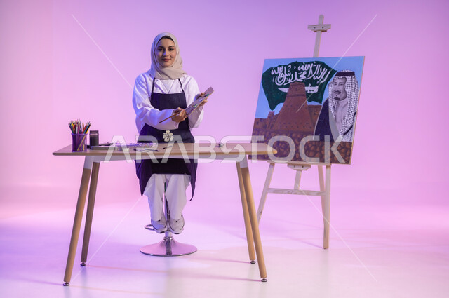 الجلوس خلف الطاولة وبجانبها لوحة معالم وعلم المملكة، النظر للكاميرا بإيماءات السعادة والسرور، بورتريه لرسامة عربية خليجية سعودية ترتدي اللباس الكاجوال ومريلة الرسم، استخدام أدوات ومعدات التلوين، استذكار اليوم الوطني السعودي، الجسم بالطول الكامل، خلفية ملونة