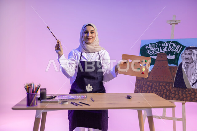 النظر للكاميرا بإيماءات السعادة والسرور، بورتريه لرسامة عربية خليجية سعودية ترتدي اللباس الكاجوال ومريلة الرسم، استخدام أدوات ومعدات التلوين، الجلوس خلف الطاولة وبجانبها لوحة معالم وعلم المملكة، استذكار اليوم الوطني السعودي، أنشطة ترفيهية فنية ممتعة، خلفية ملونة