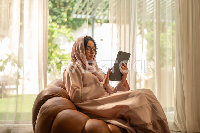 مراقبة مواقع التواصل الاجتماعي، تطور الأجهزة والتقنيات الحديثة, إنجاز مهام العمل عن بعد, مشاهدة الأفلام والمسلسلات المفضلة، البحث عن شيء ما عبر الانترنت، امرأة عربية خليجية سعودية تجلس على أريكة في غرفة المعيشة و تمسك بيدها جهاز التابليت