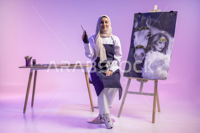النظر للكاميرا بإيماءات السعادة والسرور، استخدام ريشة التلوين والرسم، الجلوس أمام لوحة خيل عربي أصيل أبيض وبجانبه إمرأة، عشق الحصان السعودي الأصيل، بورتريه لرسامة عربية خليجية سعودية ترتدي اللباس الكاجوال ومريلة الرسم، الجسم بالطول الكامل، خلفية ملونة