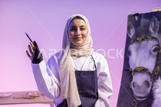 استخدام ريشة التلوين والرسم، الجلوس أمام لوحة خيل عربي أصيل أبيض وبجانبه إمرأة، عشق الحصان السعودي الأصيل، بورتريه مقرب لرسامة عربية خليجية سعودية ترتدي اللباس الكاجوال ومريلة الرسم، النظر للكاميرا بإيماءات السعادة والسرور، خلفية ملونة