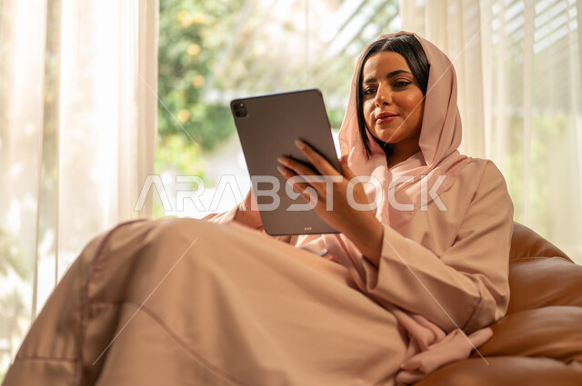 تصفح مواقع التواصل الاجتماعي، التسوق الالكتروني عبر الأونلاين، استخدام الأجهزة والتقنيات الحديثة والمتطورة, إنجاز مهام العمل عن بعد, التطبيقات التقنية للأجهزة اللوحية, امرأة عربية خليجية سعودية تجلس على أريكة في غرفة المعيشة و تمسك بيدها جهاز التابليت