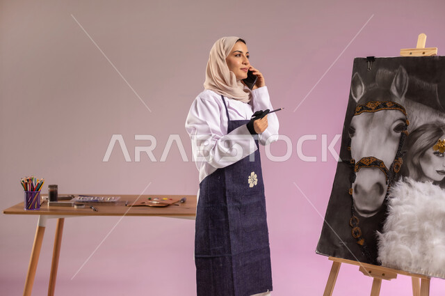 استخدام التقنيات الحديثة، إجراء مكالمة هاتفية، توارث حب الجياد من الأجداد، بورتريه لرسامة عربية خليجية سعودية ترتدي اللباس الكاجوال ومريلة الرسم، استخدام ريشة التلوين والرسم، الوقوف أمام لوحة خيل عربي أصيل أبيض وبجانبه إمرأة، خلفية ملونة