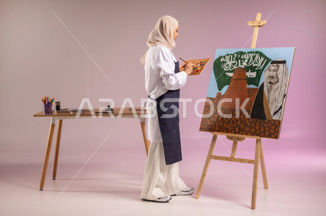 إحياء ذكرى اليوم الوطني السعودي، استخدام أدوات ومعدات التلوين، المناسبات والأعياد الوطنية، الوقوف أمام لوحة معالم وعلم المملكة، بورتريه لرسامة عربية خليجية سعودية ترتدي اللباس الكاجوال ومريلة الرسم، أنشطة ترفيهية فنية، ممارسة الرسم والفنون اليدوية، خلفية ملونة