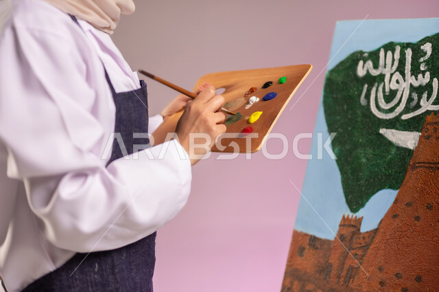 استخدام أدوات ومعدات التلوين، المناسبات والأعياد الوطنية، الوقوف أمام لوحة معالم وعلم المملكة، بورتريه لرسامة عربية خليجية سعودية ترتدي اللباس الكاجوال ومريلة الرسم، أنشطة ترفيهية فنية ممتعة، ممارسة الرسم والفنون اليدوية، استذكار اليوم الوطني، خلفية ملونة
