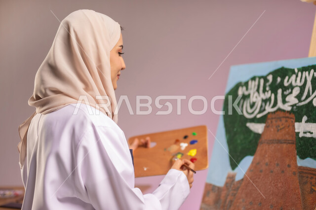 الجلوس أمام لوحة معالم وعلم المملكة، بورتريه مقرب لرسامة عربية خليجية سعودية ترتدي اللباس الكاجوال ومريلة الرسم، أنشطة ترفيهية فنية ممتعة، ممارسة الرسم والفنون اليدوية، استذكار اليوم الوطني السعودي، استخدام أدوات ومعدات التلوين، خلفية ملونة