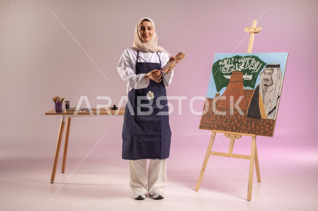ممارسة الرسم والفنون اليدوية، استذكار اليوم الوطني، استخدام أدوات ومعدات التلوين، الوقوف أمام لوحة معالم وعلم المملكة، النظر للكاميرا بإيماءات السعادة، بورتريه لرسامة عربية خليجية سعودية ترتدي اللباس الكاجوال ومريلة الرسم، الجسم بالطول الكامل، خلفية ملونة