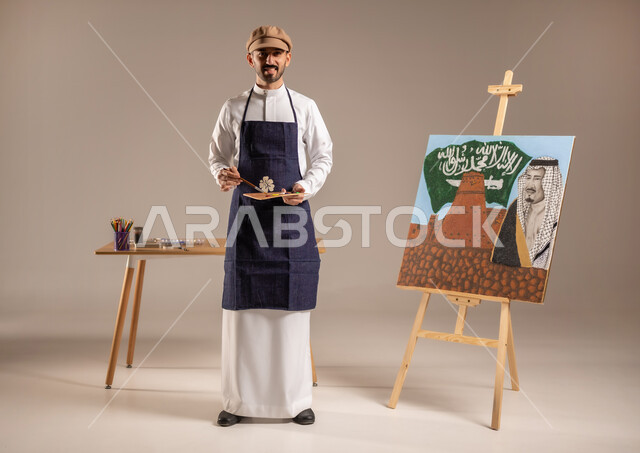 استخدام أدوات ومعدات التلوين، الوقوف أمام لوحة معالم وعلم المملكة، النظر للكاميرا بإيماءات السعادة، بورتريه لرسام عربي خليجي سعودي يرتدي الثوب التقليدي ومريلة الرسم، إحياء ذكرى اليوم الوطني، ممارسة هواية الرسم والفنون اليدوية، الجسم بالطول الكامل، خلفية ملونة