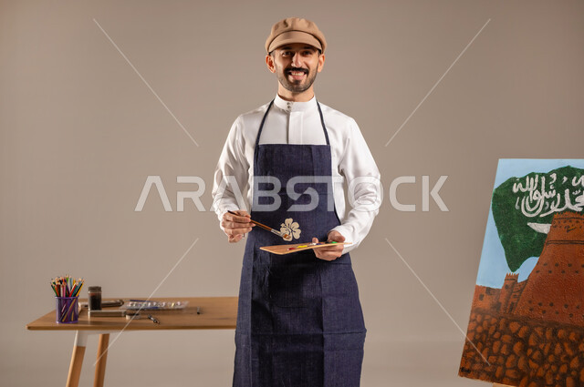 Enjoyable artistic recreational activities, standing in front of a painting of the landmarks and flag of the Kingdom, looking at the camera with happy gestures, a portrait of a Saudi Gulf Arab painter wearing a traditional dress and a drawing apron with drawing and coloring equipment in his hand, commemorating the National Day, practicing the hobby of drawing and hand arts, a colorful background.