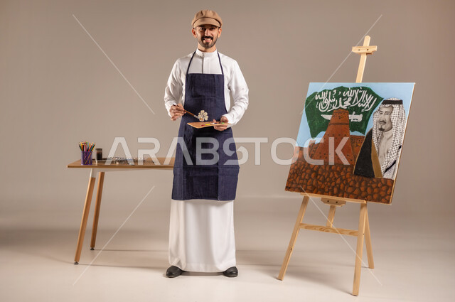 الوقوف أمام لوحة معالم وعلم المملكة، النظر للكاميرا بإيماءات السعادة، بورتريه لرسام عربي خليجي سعودي يرتدي الثوب التقليدي ومريلة الرسم وبيده معدات الرسم والتلوين، ممارسة هواية الرسم والفنون اليدوية، إحياء ذكرى اليوم الوطني، الجسم بالطول الكامل، خلفية ملونة