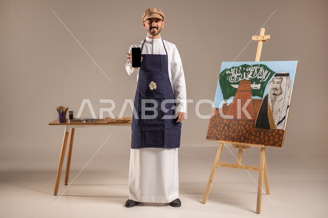 النظر للكاميرا بإيماءات السعادة، الرسم والفنون اليدوية، بورتريه لرسام عربي خليجي سعودي يرتدي الثوب التقليدي ومريلة الرسم، الوقوف أمام لوحة معالم وعلم المملكة، استخدام التقنيات الحديثة، شاشة سوداء فارغة، إحياء ذكرى اليوم الوطني، الجسم بالطول الكامل، خلفية ملونة