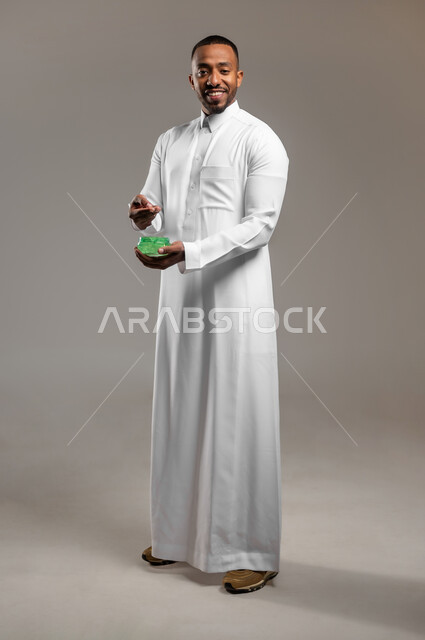 ترطيب وتنظيف وتفتيح الجلد، النظر الى الكاميرا بإيماءات السعادة والسرور، بورتريه لشاب عربي خليجي سعودي يرتدي الثوب التقليدي يضع كريم المرطب على الوجه، مفهوم العناية والإهتمام بنضارة وتحسين البشرة، استخدام مكونات تجميلية شبابية، الجسم بالطول الكامل، خلفية رمادية