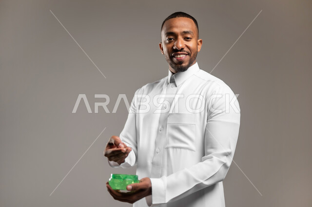 النظر الى الكاميرا بإيماءات السعادة والسرور، ترطيب وتنظيف وتفتيح الجلد، بورتريه مقرب لشاب عربي خليجي سعودي يرتدي الثوب التقليدي يضع كريم المرطب على الوجه، مفهوم العناية والإهتمام بنضارة وتحسين البشرة، استخدام مكونات تجميلية شبابية، خلفية رمادية