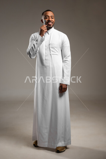 الاهتمام بالمظهر الخارجي، النظر الى الكاميرا بإيماءات الفرح والسرور، المحافظة على الأناقة والنظافة الشخصية، بورتريه لرجل عربي خليجي سعودي يرتدي الثوب التقليدي، القيام بإزالة شعر الوجه باستخدام شفرة الحلاقة الرجالية، الجسم بالطول الكامل، خلفية رمادية