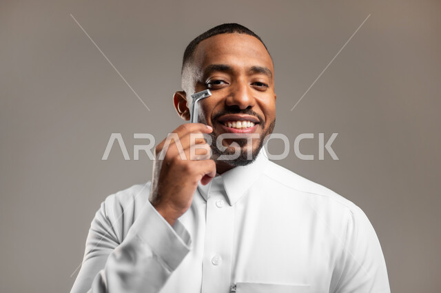 النظر الى الكاميرا بإيماءات الفرح والسرور، المحافظة على الأناقة والنظافة الشخصية، مفهوم العناية بالبشرة والاهتمام بالمظهر الخارجي، بورتريه مقرب لرجل عربي خليجي سعودي يرتدي الثوب التقليدي، القيام بإزالة شعر الوجه باستخدام شفرة الحلاقة الرجالية، خلفية رمادية