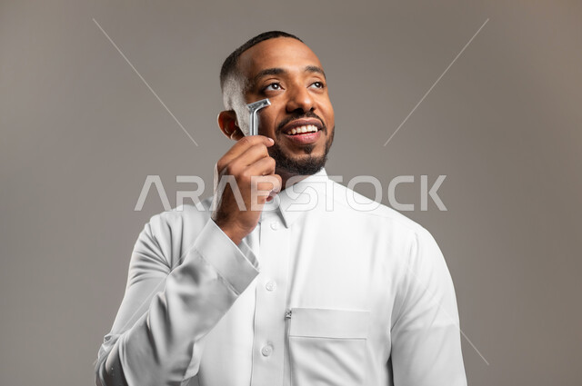 المحافظة على الأناقة والنظافة الشخصية، مفهوم العناية بالبشرة والاهتمام بالمظهر الخارجي، بورتريه مقرب لرجل عربي خليجي سعودي يرتدي الثوب التقليدي، القيام بإزالة شعر الوجه باستخدام شفرة الحلاقة الرجالية، خلفية رمادية