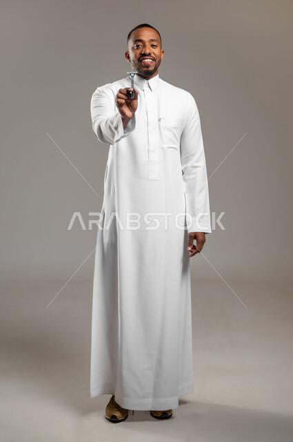 الاهتمام بالمظهر الخارجي والعناية بالبشرة، الحفاظ على الأناقة والنظافة الشخصية، النظر الى الكاميرا بإيماءات السعادة والسرور، بورتريه لرجل عربي خليجي سعودي يرتدي الثوب التقليدي يمسك بيده شفرة الحلاقة الرجالية، الجسم بالطول الكامل، خلفية رمادية