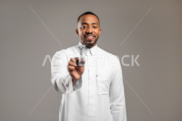 Maintaining elegance and personal hygiene, looking at the camera with gestures of happiness and pleasure, taking care of the skin and taking care of the external appearance, a close-up portrait of a Saudi Gulf Arab man wearing a traditional thobe, holding a men’s razor in his hand, gray background.