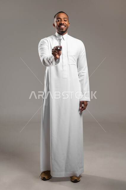 النظر الى الكاميرا بإيماءات السعادة والسرور، العناية بالبشرة والاهتمام بالمظهر الخارجي، الحفاظ على الأناقة والنظافة الشخصية، بورتريه لرجل عربي خليجي سعودي يرتدي الثوب التقليدي يمسك بيده شفرة الحلاقة الرجالية، الجسم بالطول الكامل، خلفية رمادية