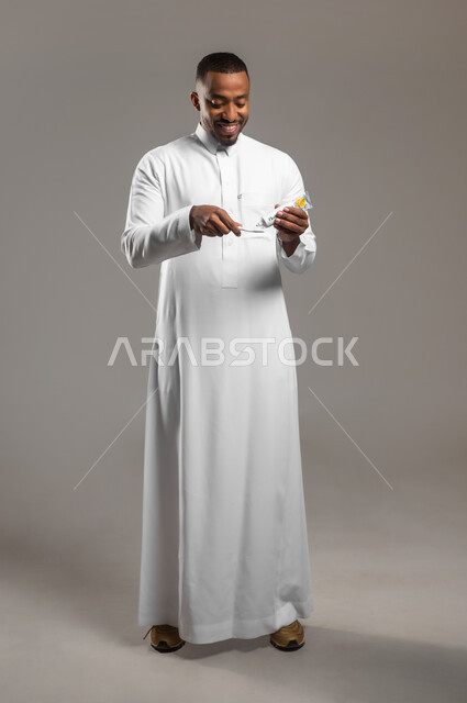 تجنب التسوس وأمراض اللثة، مفهوم العناية والإهتمام بصحة الأسنان، روتين نظافة الفم اليومية، بورتريه لرجل عربي خليجي سعودي يرتدي الثوب التقليدي يقوم بتنظيف أسنانه باستخدام الفرشاة والمعجون، الجسم بالطول الكامل، خلفية رمادية
