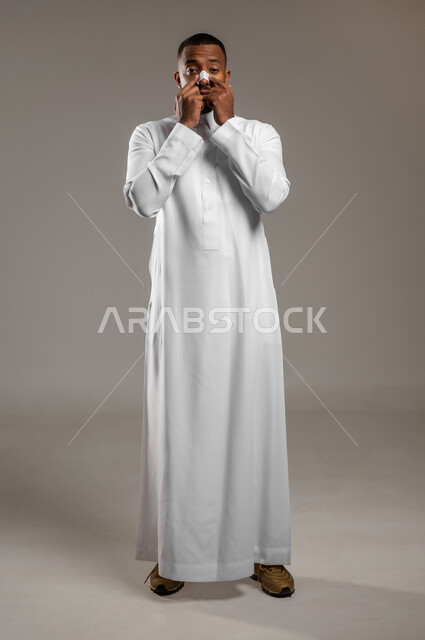 Caring for the freshness and improvement of the skin, youthful cosmetic ingredients, using a pore-cleaning mask, portrait of a Saudi Gulf Arab young man wearing a traditional dress, applying nose patches to get rid of blackheads and dead skin, looking at the camera with astonished gestures, full-length body, gray background.