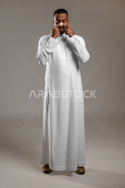 Youthful cosmetic ingredients, using a pore-cleaning mask, portrait of a handsome young Saudi Arabian Gulf man wearing a traditional dress, applying nose patches to get rid of blackheads and dead skin, the concept of care and concern for the freshness and improvement of the skin, full-length body, gray background