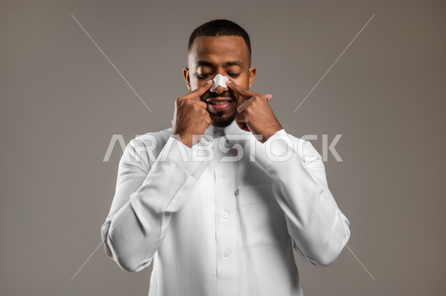 استخدام ماسك تنظيف المسام، بورتريه مقرب لشاب وسيم عربي خليجي سعودي يرتدي الثوب التقليدي يضع لصقات الأنف للتخلص من الرؤوس السوداء والجلد الميت، مفهوم العناية والإهتمام بنضارة وتحسين البشرة، مكونات تجميلية شبابية، خلفية رمادية