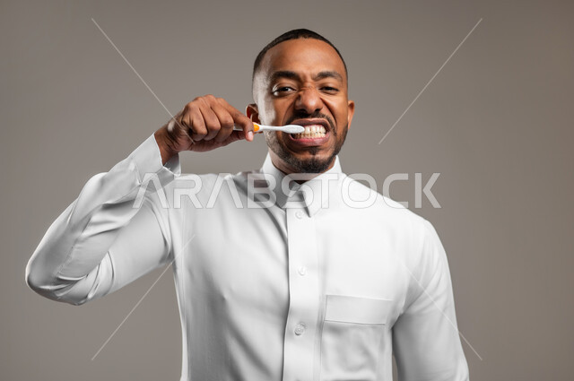 النظر الى الكاميرا بإيماءات السعادة والسرور، روتين نظافة الفم اليومية، بورتريه مقرب لرجل عربي خليجي سعودي يرتدي الثوب التقليدي يقوم بتنظيف أسنانه باستخدام الفرشاة، مفهوم العناية والإهتمام بصحة الأسنان، تجنب التسوس وأمراض اللثة، خلفية رمادية