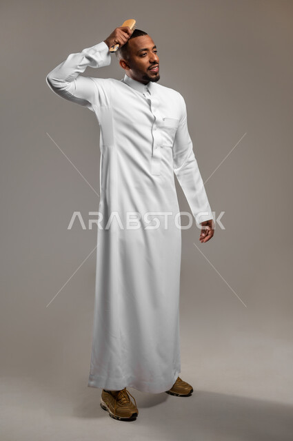 مفهوم الأناقة والمظهر الخارجي والعناية والاهتمام بالجمال، تصفيف وترتيب الشعر، بورتريه لرجل عربي خليجي سعودي يرتدي الثوب التقليدي يقوم بتمشيط شعره باستخدام فرشاة الشعر ( المشط )، الجسم بالطول الكامل، خلفية رمادية