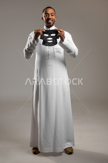 مفهوم العناية والاهتمام بالمظهر الخارجي، استخدام مكونات تجميلية، ماسك تنظيف وتقشير البشرة، بورتريه لشاب عربي خليجي سعودي يرتدي الثوب التقليدي يقوم بنزع  قناع الفحم الاسود من على وجهه للتخلص من الرؤوس السوداء، الجسم بالطول الكامل، خلفية رمادية