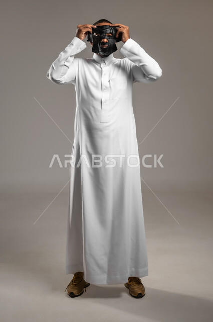 استخدام مكونات تجميلية، ماسك تنظيف وتقشير البشرة، بورتريه لشاب وسيم عربي خليجي سعودي يرتدي الثوب التقليدي يقوم بوضع قناع الفحم الاسود على وجهه للتخلص من الرؤوس السوداء، مفهوم العناية والاهتمام بالمظهر الخارجي، الجسم بالطول الكامل، خلفية رمادية