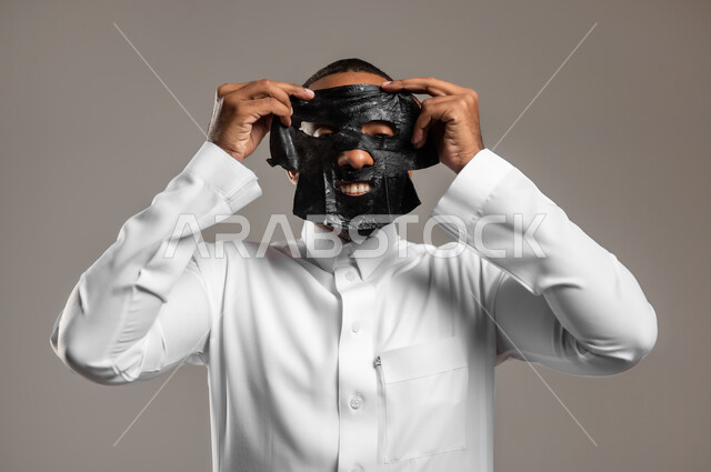 ماسك تنظيف وتقشير البشرة، استخدام مكونات تجميلية، بورتريه مقرب لشاب وسيم عربي خليجي سعودي يرتدي الثوب التقليدي يقوم بوضع قناع الفحم الاسود على وجهه للتخلص من الرؤوس السوداء، مفهوم العناية والاهتمام بالمظهر الخارجي، خلفية رمادية