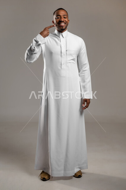 الحصول على إبتسامة هوليود باستخدام قشور فينير من الخزف، تجميل وتبييض الأسنان، بورتريه لرجل عربي خليجي سعودي يرتدي الثوب التقليدي يشير بإصبع السبابة على أسنانه البيضاء، مفهوم العناية والإهتمام بالصحة والمظهر الخارجي ، الجسم بالطول الكامل، خلفية رمادية