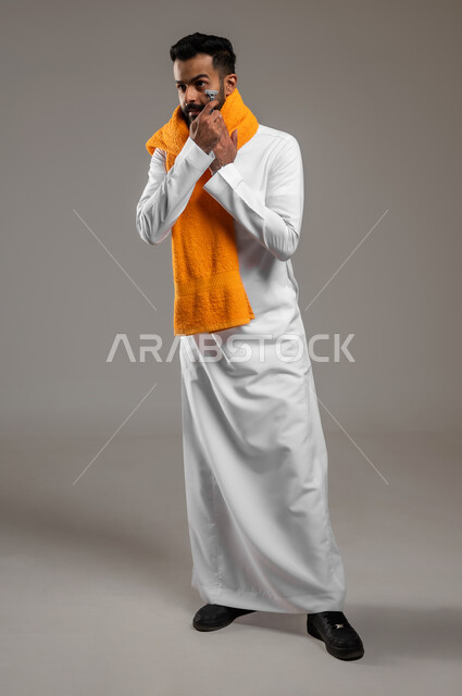 الاهتمام بالمظهر الخارجي ومفهوم العناية بالبشرة، المحافظة على الأناقة والنظافة الشخصية، بورتريه لرجل عربي خليجي سعودي يرتدي الثوب التقليدي يضع المنشفة البرتقالية على عنقه ويقوم بإزالة شعر الوجه باستخدام شفرة الحلاقة الرجالية، الجسم بالطول الكامل، خلفية رمادية