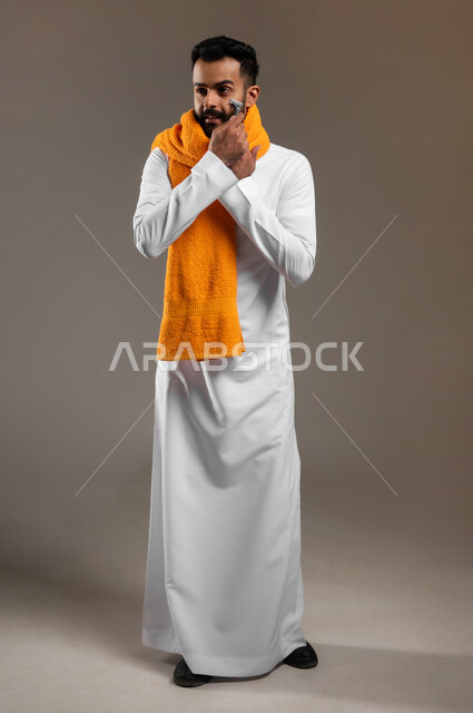 المحافظة على الأناقة والنظافة الشخصية، مفهوم العناية بالبشرة والاهتمام بالمظهر الخارجي، بورتريه لرجل عربي خليجي سعودي يرتدي الثوب التقليدي يضع المنشفة البرتقالية على عنقه ويقوم بإزالة شعر الوجه باستخدام شفرة الحلاقة الرجالية، الجسم بالطول الكامل، خلفية رمادية