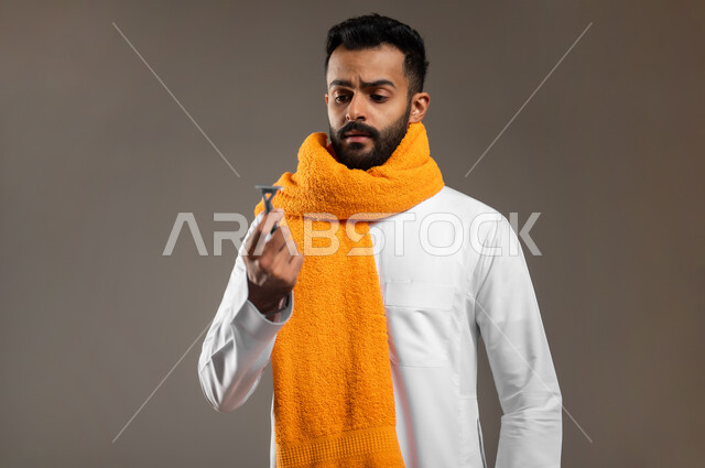 الحفاظ على الأناقة والنظافة الشخصية، العناية بالبشرة والاهتمام بالمظهر الخارجي، بورتريه مقرب لرجل عربي خليجي سعودي يرتدي الثوب التقليدي يضع المنشفة البرتقالية على عنقه، النظر الى شفرة الحلاقة الرجالية بتعابير الاستغراب، خلفية رمادية